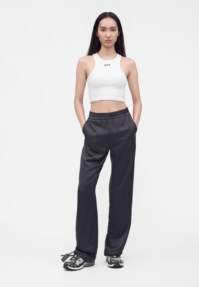 Femme aux longs cheveux noirs portant un crop top blanc avec le texte "OFF", un pantalon noir ample et des baskets noires et blanches, debout les mains dans les poches.