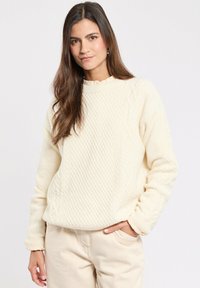 Maglione lavorato a maglia color crema con design a treccia testurizzata, scollatura ampia e polsini a costine, abbinato a pantaloni beige chiaro.