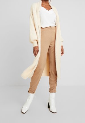 Femme portant un haut blanc à bords festonnés, un pantalon jogger beige, un long cardigan crème et des bottines blanches à talons, debout devant un fond gris.