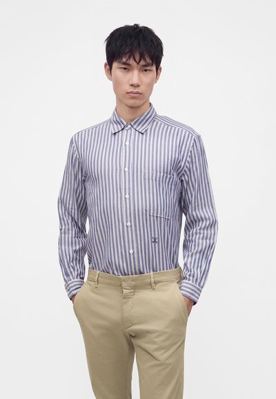 Jeune homme portant une chemise à rayures bleues et blanches rentrée dans un pantalon beige, les mains dans les poches, debout devant un fond uni.