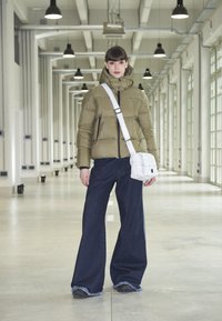 Olivový puffrák s vysokým golierom, skombinovaný s širokými tmavými denimovými džínsami. Biela crossbody taška má prešívaný dizajn a čierne akcenty.