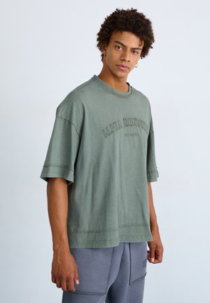 Jeune homme aux cheveux bouclés portant un t-shirt vert surdimensionné Alpha Industries et un pantalon gris, debout devant un fond clair uni.