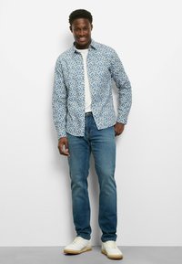 Weißes Hemd unter einem blau geblümten Hemd mit Knöpfen, kombiniert mit hellblauen Jeans und weißen Sneakern mit Gummisohlen.