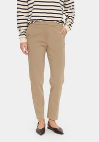 Pantalons fuselés beiges avec une texture lisse, dotés de poches latérales et d'un design à l'avant plat. Associés à une chemise rayée à manches longues.