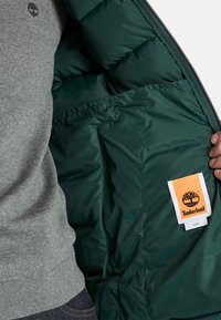 Osoba v šedém svetru drží otevřenou zelenou péřovou bundu, na které je viditelné logo Timberland a štítek s pokyny k údržbě uvnitř kapsy.