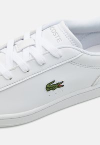 Hvit lær sneaker med en grønn brodert krokodillelogo, flate lisser og en strukturert tunge. Minimalistisk design med subtile sømdetaljer.