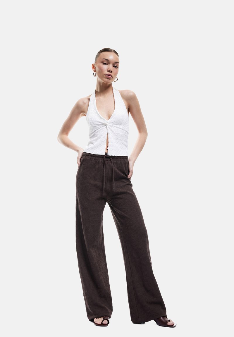 Top blanco tipo halter con un frente retorcido, combinado con pantalones de pierna ancha de textura suelta en color marrón. Sandalias negras de tacón completan el conjunto.