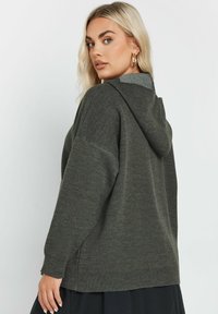 Mörkgrön huvtröja med en texturerad tyg, med en avslappnad passform, ribbade ärmslut och nederkant, som visar en sydd detalj vid halsringningen.