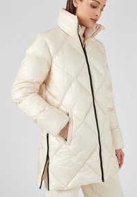 Chaqueta de plumas acolchada de color beige claro con un alto cuello, cremallera negra y bolsillos laterales. La tela tiene un acabado liso y brillante.