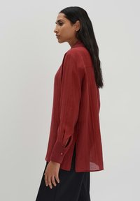 Blusa rossa a maniche lunghe con una texture leggera, fessure laterali e polsini con bottoni. Presenta un colletto e una vestibilità comoda.
