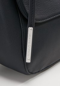 Coin d'un sac en cuir noir avec des coutures visibles et une étiquette en métal argenté gravée de "Marc O'Polo" attachée à une lanière en cuir.