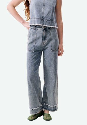 Femme portant un pantalon large en jean bleu clair et un haut court en jean avec bords effilochés, assortis à des chaussures à enfiler en filet vert.