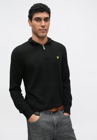 Fekete cipzáras pulóver magas gallérral, a mellkason egy kis sárga logóval; szürke farmerrel és barna övvel viselve.