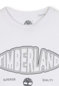T-shirt bianco in cotone con un graffito nero che riporta la scritta "TIMBERLAND" accompagnata da un logo ad albero e il testo "SUPERIOR QUALITY" sottostante.