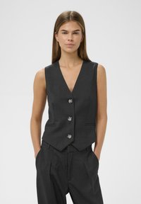 Chaleco gris a medida con escote en V profundo, cuatro botones plateados y bolsillos laterales, combinado con pantalones gris a juego. Textura suave y diseño ajustado.