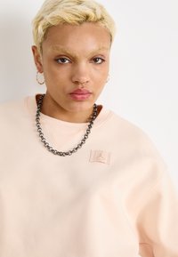 Svetloružová mikina s textured látkou, s malou nášivkou loga a jednoduchým výstrihom. Doplnená hrubým strieborným náhrdelníkom.