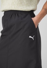 Main d'une personne dans la poche d'un short Puma noir à taille élastique, pull vert clair texturé visible, tatouage sur l'avant-bras.