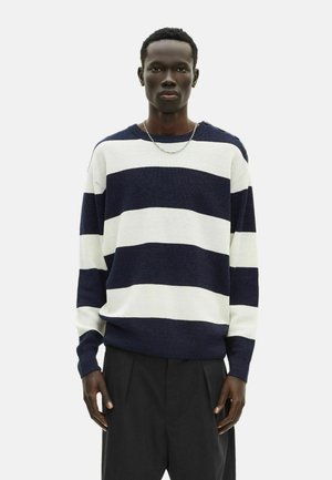 Maglione a righe navy e bianche in maglia con scollo rotondo e maniche lunghe, caratterizzato da una vestibilità ampia e polsini a coste. Pantaloni neri completano l'outfit.