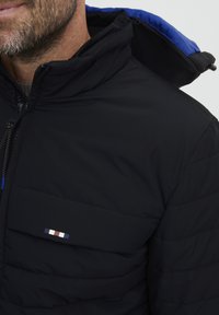 Črna podložena jakna z visokim ovratnikom in modrim notranjim podložno. Ima zadrgo in majhen večbarven logo na prsih.