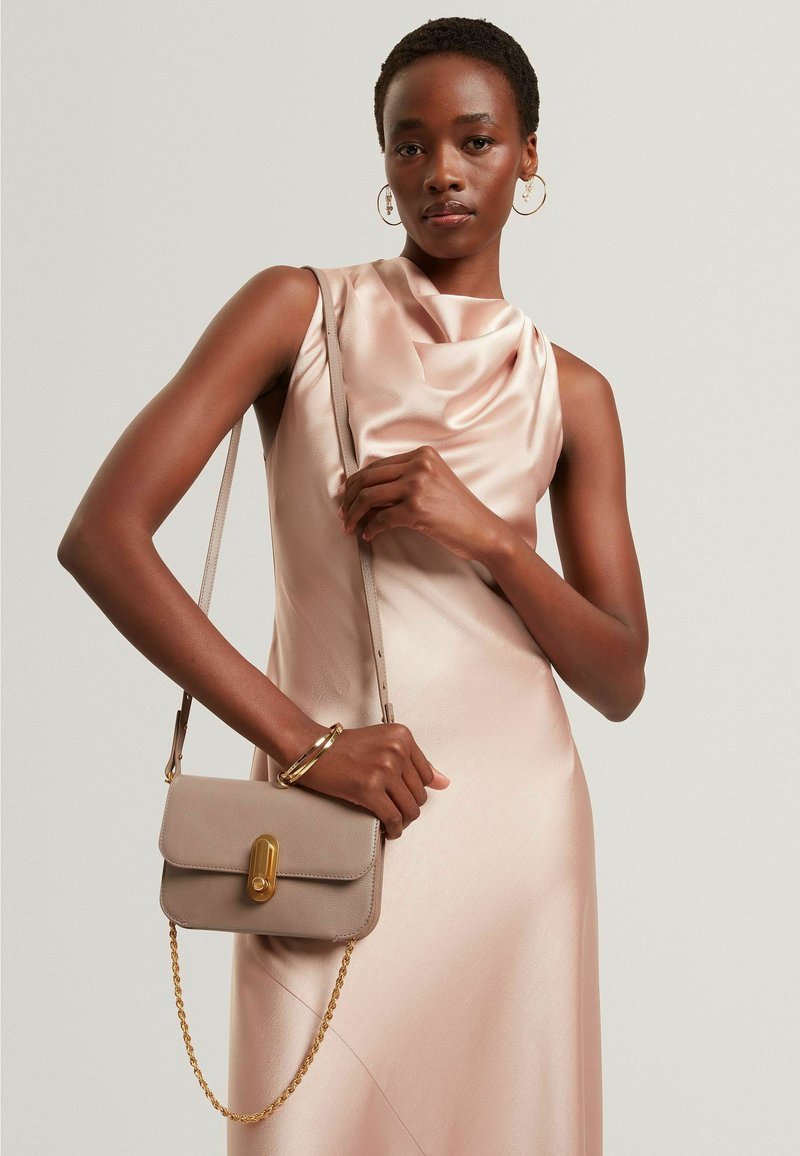 Bolso de mano de cuero beige con un broche dorado y una correa de cadena, sostenido por una modelo que lleva un vestido de satén rosa con un escote drapeado.