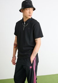 Polo nero con tessuto testurizzato e colletto con zip, abbinato a pantaloni sportivi neri con strisce rosa sui lati e un piccolo logo.