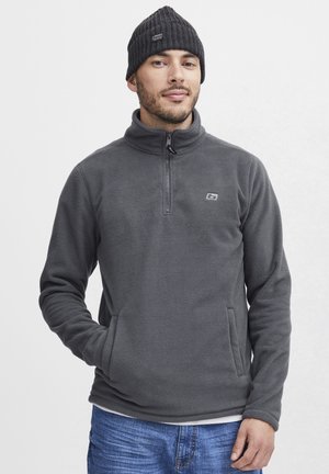 Pull en polaire gris avec un col montant et une fermeture éclair sur le haut. Il dispose de poches latérales et d'un petit logo sur la poitrine. Porté avec un bonnet noir.