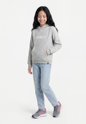 Chica con cabello largo y oscuro que lleva una sudadera con capucha gris "TERREX", vaqueros azul claro y zapatillas grises con detalles rosas, de pie y sonriendo.