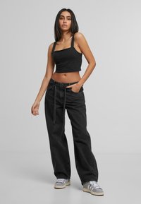 Nainen, jolla on päällään musta lyhyt tanktopaita, löysät mustat farkut vyöllä ja harmaat lenkkarit, seisomassa yksinkertaista harmaata taustaa vasten.