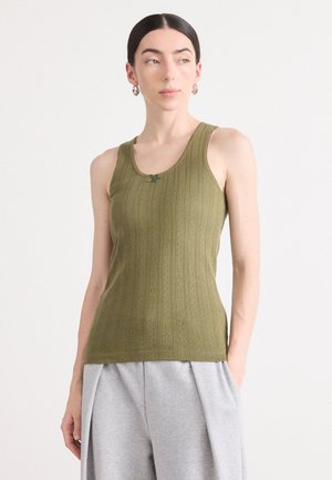 Débardeur côtelé vert olive avec un col rond et un accent décoratif au niveau de la poitrine. Porté avec un short plissé gris clair.
