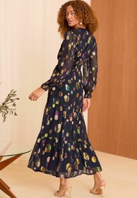Rochie lungă de culoare bleumarin, realizată din țesătură transparentă, cu buline metalice colorate, mâneci lungi, tiv în straturi, asortată cu sandale cu barete nude.
