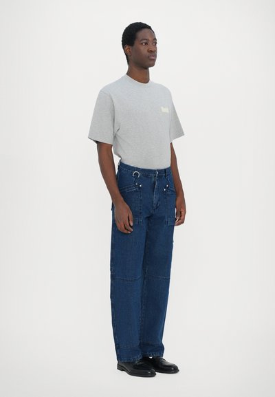 Camiseta gris de manga corta con un pequeño logo, combinada con pantalones de mezclilla azules holgados que presentan múltiples bolsillos y detalles en metal.