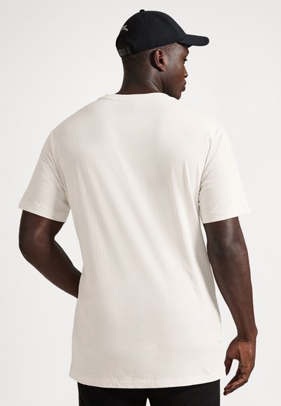 Svart mann iført en enkel hvit T-skjorte med korte ermer og en svart baseballcap, vendt bort fra kameraet mot en ensfarget hvit bakgrunn.