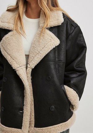 Woman wearing black leather jacket with beige shearling collar, cuffs, and hem over a white top.