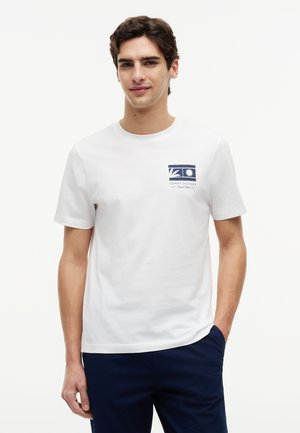 Ung mand med kort mørkt hår iført en hvid T-shirt med en lille blå grafik og marineblå bukser, stående med den ene hånd i lommen.