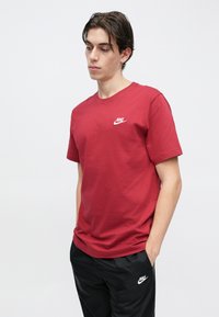 Mladi moški s temnimi lasmi, oblečen v rdečo majico Nike in črne hlače Nike, stoji z rokami v žepih pred enobarvnim ozadjem.