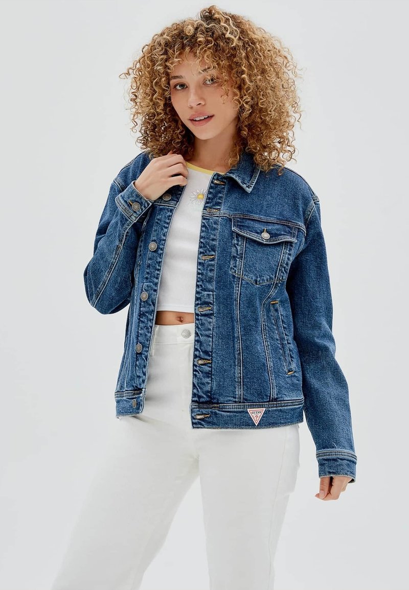 Giubbotto di jeans di colore blu scuro, con chiusura a bottoni, due tasche sul petto e una patch a forma di triangolo sull'orlo. Abbinato a una maglietta bianca e pantaloni chiari.