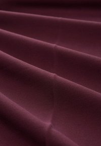 Temno burgundno blago, gladke teksture, enakomerno zloženo z vidnimi šivi in subtilnim leskom, ki prikazuje mehko, fleksibilno material.