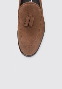Zapato de ante marrón tipo slip-on con punta redondeada y detalle de borlas en el empeine; cuenta con una suela oscura y suave y costuras en contraste.