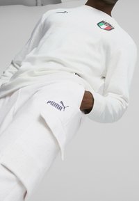 Felpa bianca con emblema della bandiera italiana e texture leggera a macchie, abbinata a pantaloni della tuta bianchi con logo Puma in viola.
