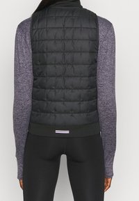 Černá prošívaná vesta s vysokým límcem, spárovaná s fialovým tričkem s dlouhým rukávem. Spodní část má černý pas s tenkým bílým detailem.