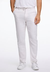 Pantalones de lino blancos con corte recto, cintura con cordón y bolsillos laterales, combinados con zapatillas blancas sobre un fondo claro.