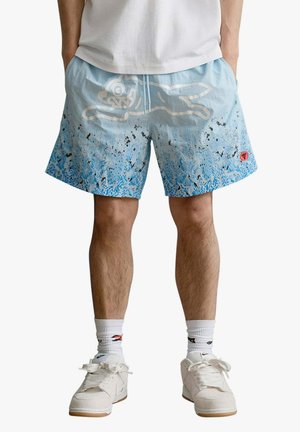 Homme portant un short bleu clair avec des éclaboussures noires et bleues, un grand imprimé animé blanc, des baskets blanches et des chaussettes blanches avec des logos.
