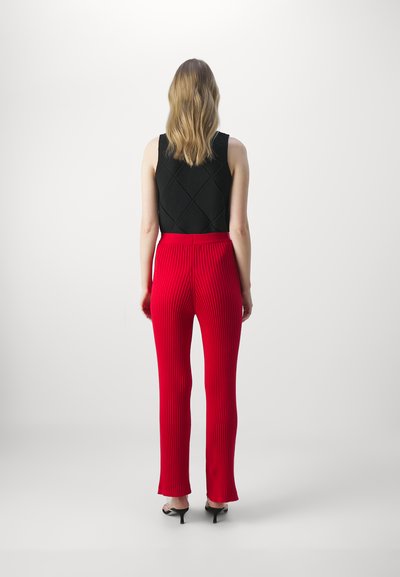 Schwarze Strickweste mit Rautenmuster, kombiniert mit hochtaillierten, roten Ripp-Hosen, die am Saum leicht ausgestellt sind, sowie schwarzen High-Heels.