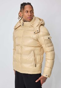 Homme aux cheveux tressés portant une doudoune à capuche beige et un pantalon noir, regardant sur le côté contre un fond clair uni.