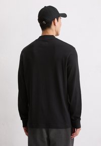 Schwarzes Langarmshirt aus weicher Baumwolle mit rundem Kragen und gerippten Bündchen, getragen mit grauen gestreiften Hosen und einer schwarzen Mütze.
