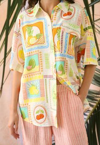 Chemise boutonnée ample avec des motifs fruités colorés portée sur un pantalon rayé rose et blanc, une main dans la poche devant un fond de plantes tropicales.