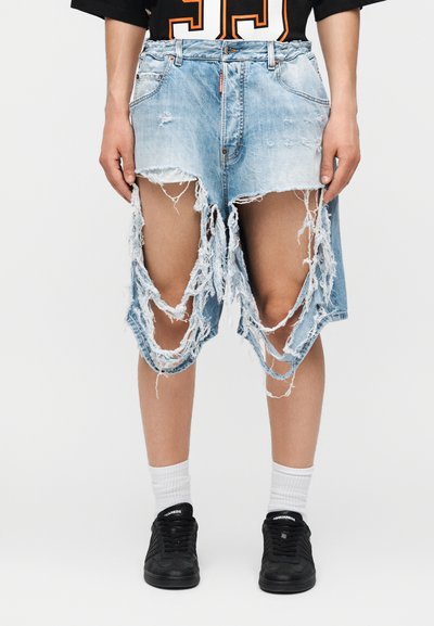 Personne portant un short en jean bleu clair très usé et déchiré, associé à des chaussettes blanches et des baskets noires.