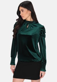 Smaragdgrünes Samtbluse mit hohem Kragen und Schlüssellochdetail, ausgestattet mit gefältelten Puffärmeln und gesmockten Manschetten. Getragen mit einem schwarzen Rock.