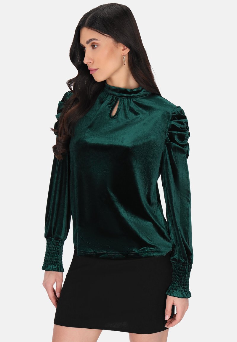 Smaragdgrünes Samtbluse mit hohem Kragen und Schlüssellochdetail, ausgestattet mit gefältelten Puffärmeln und gesmockten Manschetten. Getragen mit einem schwarzen Rock.