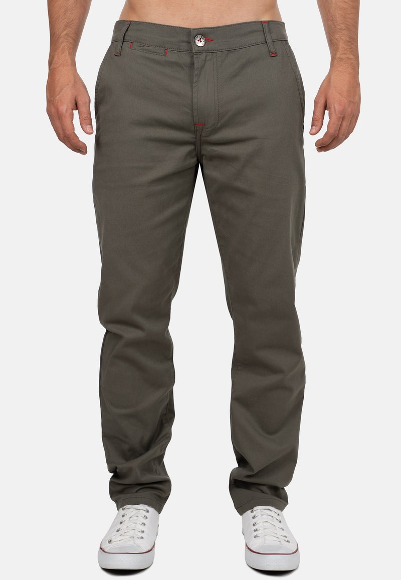 Pantalons kaki en tissu mélangé de coton. Caractéristiques de surpiqûres rouges, coupe droite et fermeture à bouton à l'avant. Portés avec des baskets blanches.